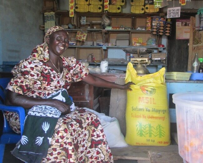 Hellen is seated in a colourful dress as she shows off her shop, full of products in sacks and plastic crates.