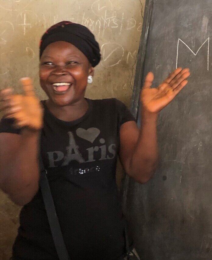 Woman in a black hat and black, 'I love Paris' t-shirt laughs and claps in front of a blackboard.