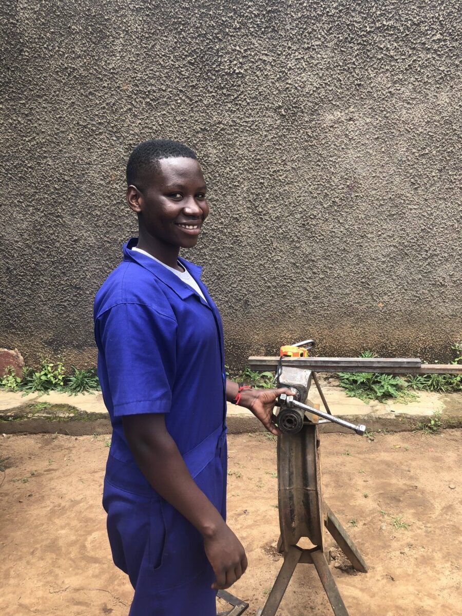 Sandra stands at a bit of machinery and smiles at the camera. She is wearing blue overalls.