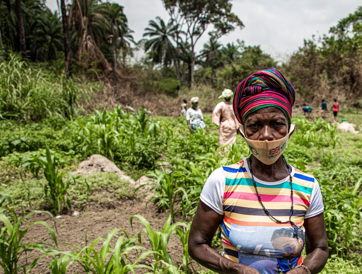 A woman stands in her farm looking at the camera and holding some crops.